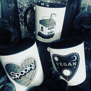 May include: Three white ceramic mugs with black rims. The mugs have different designs. One mug has a cartoon image of a milk carton with a skull and crossbones and the text "Dairy is Scary". Another mug has a black heart with a spiderweb design and the text "Spooky". The third mug has a black design of a  pendulum with the text "Vegan".