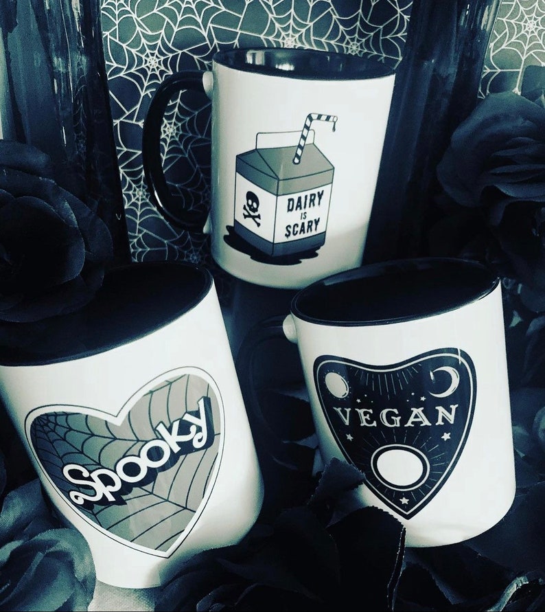May include: Three white ceramic mugs with black rims. The mugs have different designs. One mug has a cartoon image of a milk carton with a skull and crossbones and the text "Dairy is Scary". Another mug has a black heart with a spiderweb design and the text "Spooky". The third mug has a black design of a  pendulum with the text "Vegan".