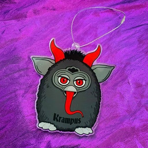 Puede incluir: Un adorno con tem&aacute;tica de Krampus que presenta una criatura de dibujos animados con ojos, cuernos y lengua rojos. El adorno tiene un cuerpo negro, detalles en gris y la palabra "Krampus" impresa. Una cuerda plateada est&aacute; unida para colgar.