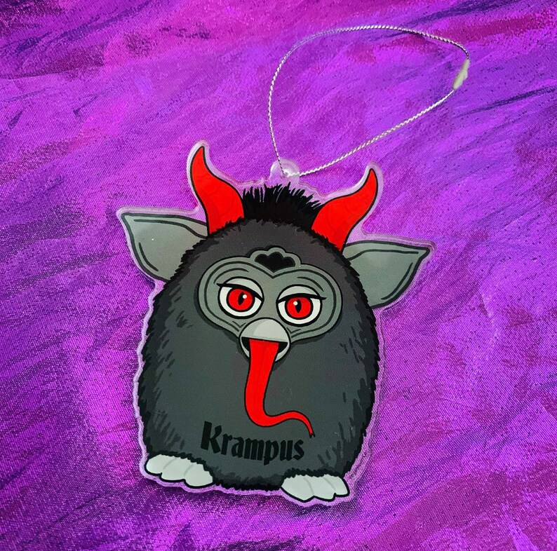 Puede incluir: Un adorno con tem&aacute;tica de Krampus que presenta una criatura de dibujos animados con ojos, cuernos y lengua rojos. El adorno tiene un cuerpo negro, detalles en gris y la palabra "Krampus" impresa. Una cuerda plateada est&aacute; unida para colgar.