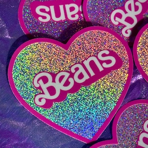 May include: A heart-shaped sticker with a holographic glitter finish featuring the word BEANS in white, bold, rounded lettering. The sticker has a thick magenta border and a reflective surface that displays a spectrum of rainbow colors.