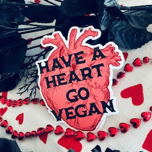 Puede incluir: Una pegatina roja de corazón anatómico con el texto "Have a heart go vegan" en letras negras.