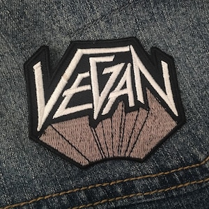 May include: A black and white embroidered patch with the word "VEGAN" in a stylized font. The patch is sewn onto a blue denim fabric.