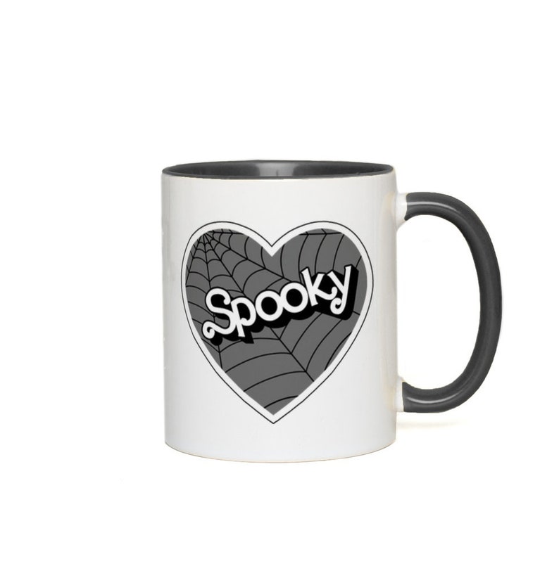 May include: A white ceramic mug with a black rim and handle. The mug features a black and white illustration of a heart with a spiderweb design and the word "Spooky" inside the heart.