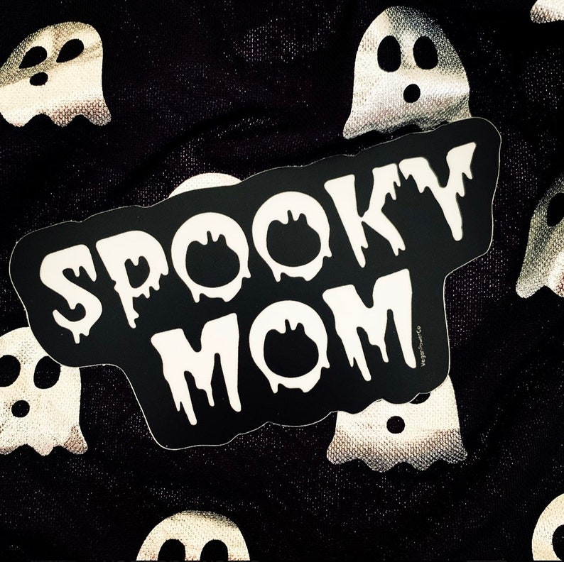 May include: Black and white sticker with the text "Spooky Mom" in dripping white letters on a black background. The sticker is on a black fabric background with white ghost shapes.