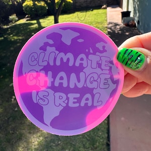 May include: A round sticker with a purple and pink background. The sticker features a white outline of the Earth and the text "Climate Change is Real" in white.