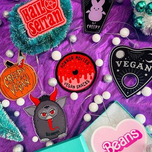Puede incluir: Una colecci&oacute;n de adornos navide&ntilde;os originales. Incluyen dise&ntilde;os con las frases "Hail Seitan", una calabaza con "Creepy Vegan", una rosquilla con "Horror Movies Vegan Snacks", un ata&uacute;d con un conejito y "Cruelty Free But Creepy", una tabla Ouija con "Vegan", Krampus y un coraz&oacute;n rosa con "Beans".