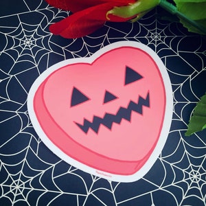 Puede incluir: Una pegatina rosa en forma de corazón con una cara de calabaza negra. La pegatina tiene un contorno blanco.