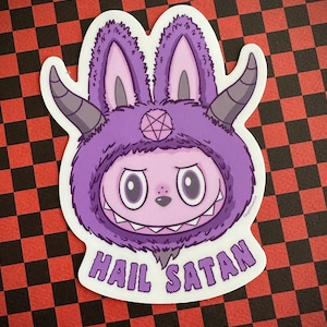 May include: A sticker featuring a purple cartoon character with bunny ears, horns, and a pentagram on its forehead. The character has a wide, toothy grin and the words "HAIL SATAN" are printed below it.