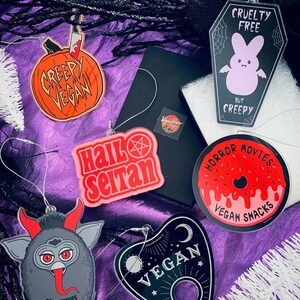 Puede incluir: Una colecci&oacute;n de adornos tem&aacute;ticos de Halloween con varios dise&ntilde;os y texto. Los adornos incluyen una calabaza con un cuchillo, un ata&uacute;d con un conejito, una rosquilla, una tabla Ouija y una criatura parecida a Krampus. El texto incluye "Creepy Vegan", "Hail Satan", "Horror Movies Vegan Snacks" y "Vegan".
