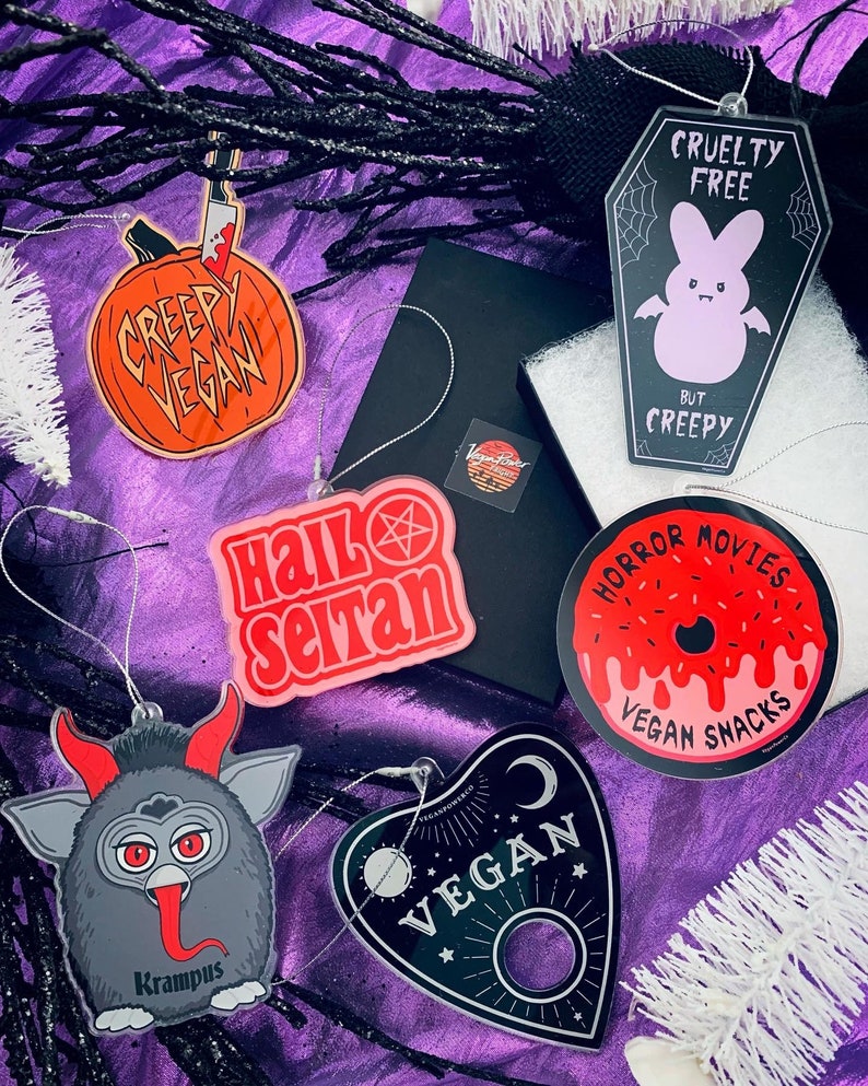 Puede incluir: Una colecci&oacute;n de adornos tem&aacute;ticos de Halloween con varios dise&ntilde;os y texto. Los adornos incluyen una calabaza con un cuchillo, un ata&uacute;d con un conejito, una rosquilla, una tabla Ouija y una criatura parecida a Krampus. El texto incluye "Creepy Vegan", "Hail Satan", "Horror Movies Vegan Snacks" y "Vegan".