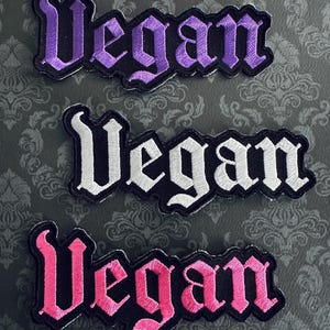May include: Three embroidered patches with the word "Vegan" in a gothic font. The patches are in purple, white, and pink, each with a black border. The background is a patterned gray fabric.