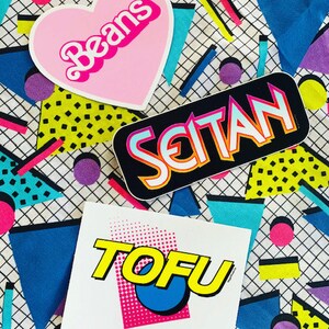 May include: Three colorful stickers with a retro 80s design. The stickers say "Beans" in a pink heart shape, "Seitan" in a black rectangle, and "TOFU" in a white rectangle with a pink, blue, and yellow design.