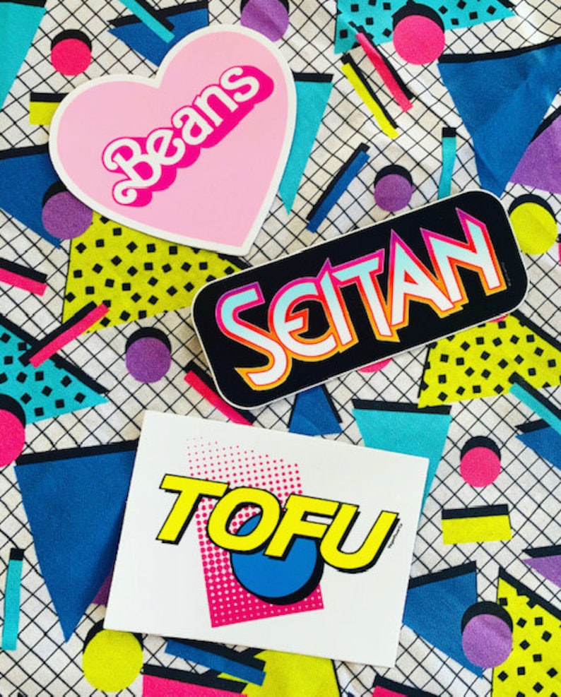 May include: Three colorful stickers with a retro 80s design. The stickers say "Beans" in a pink heart shape, "Seitan" in a black rectangle, and "TOFU" in a white rectangle with a pink, blue, and yellow design.