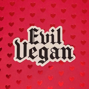 May include: A white sticker with black text that reads "Evil Vegan" in a gothic font. The sticker is on a red background with a pattern of small red hearts.