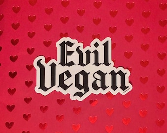 May include: A white sticker with black text that reads "Evil Vegan" in a gothic font. The sticker is on a red background with a pattern of small red hearts.