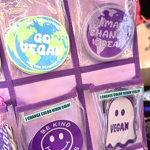 May include: A purple organizer with clear plastic pockets containing various stickers. The stickers feature different messages and designs, including "Go Vegan", "Climate Change is Real", "Be Kind to Animals", and "Vegan". Some of the stickers change color when cold.