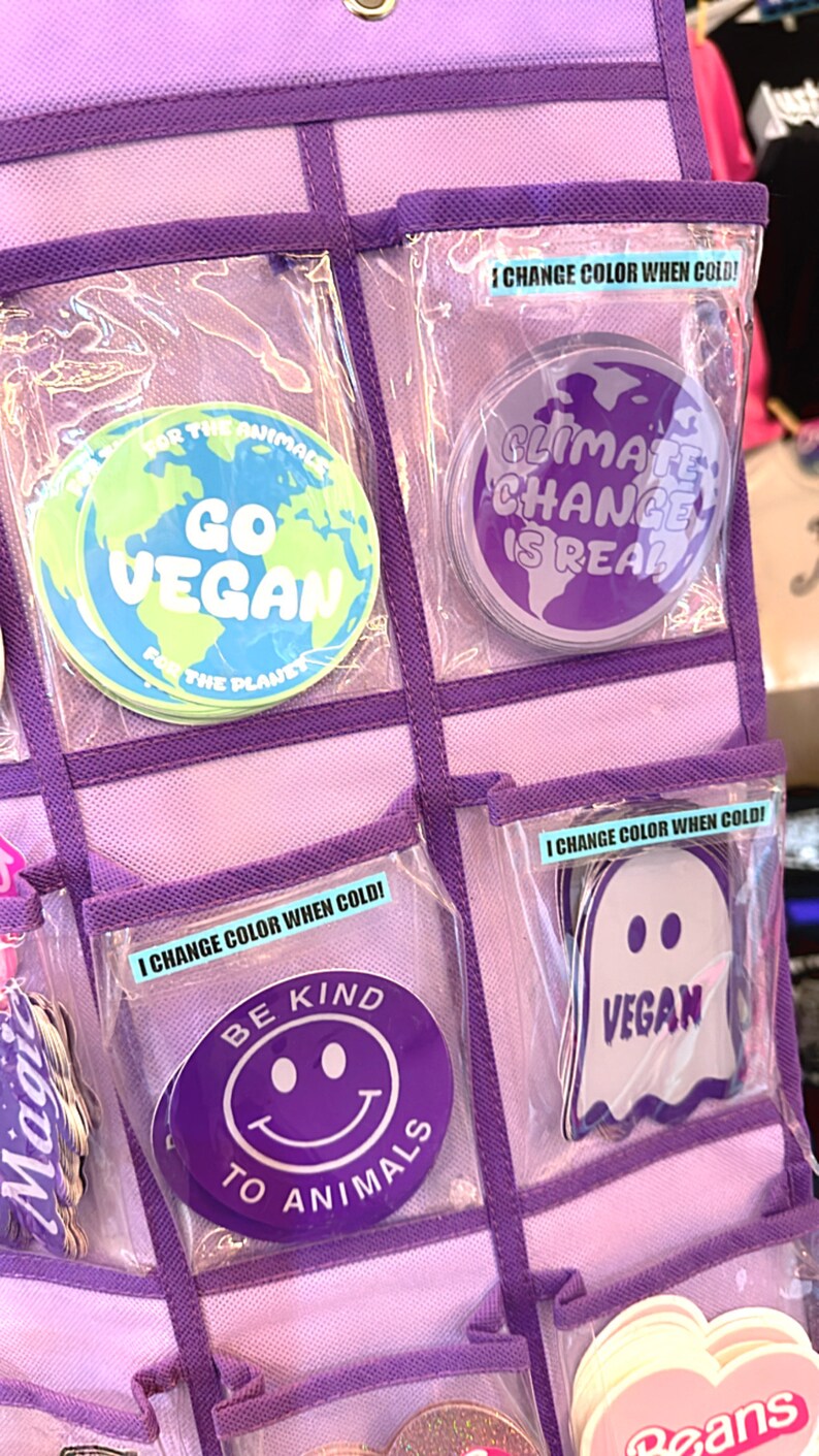 May include: A purple organizer with clear plastic pockets containing various stickers. The stickers feature different messages and designs, including "Go Vegan", "Climate Change is Real", "Be Kind to Animals", and "Vegan". Some of the stickers change color when cold.