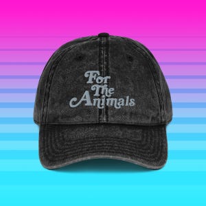 Pode incluir: Boné de beisebol preto com o texto bordado "For The Animals" em uma fonte estilizada. O boné tem uma aba curva e um visual vintage desgastado. O fundo é um gradiente de rosa, roxo e azul.