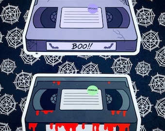 May include: Two Halloween-themed VHS tape stickers. One is gray with the word "BOO!!" and a "Spooky" sticker. The other is black with red dripping blood and a "Horror" sticker. The background is black with white spiderwebs.