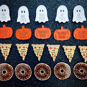 May include: A set of 16 enamel pins featuring various gender identities and vegan messages. The pins are in the shape of ghosts, pumpkins, pizza slices, and donuts. The pins are all colorful and have a glossy finish.