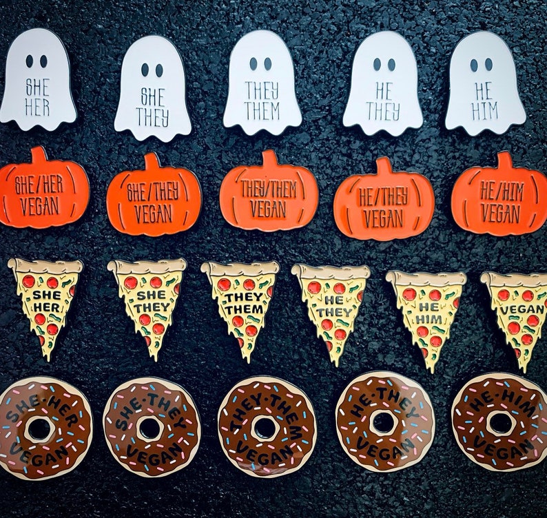 May include: A set of 16 enamel pins featuring various gender identities and vegan messages. The pins are in the shape of ghosts, pumpkins, pizza slices, and donuts. The pins are all colorful and have a glossy finish.