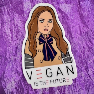 May include: A die-cut sticker featuring a cartoon illustration of a person with long brown hair, wearing a tan coat and a blue and pink bow tie. The sticker has the text "VEGAN IS THE FUTURE" in a bold font.
