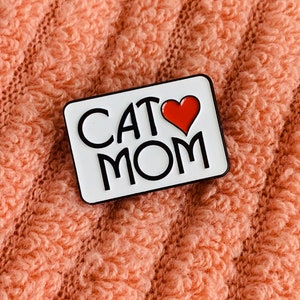 K&ouml;nnte beinhalten: Wei&szlig;er Emaille-Pin mit dem Text "Cat Mom" und einem roten Herzen.
