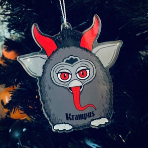 Puede incluir: Adorno navide&ntilde;o gris con tem&aacute;tica de Krampus, con cuernos, ojos y lengua rojos. El adorno tiene un dise&ntilde;o de dibujos animados, con la palabra "Krampus" impresa en negro. Cuelga de un &aacute;rbol de Navidad negro.