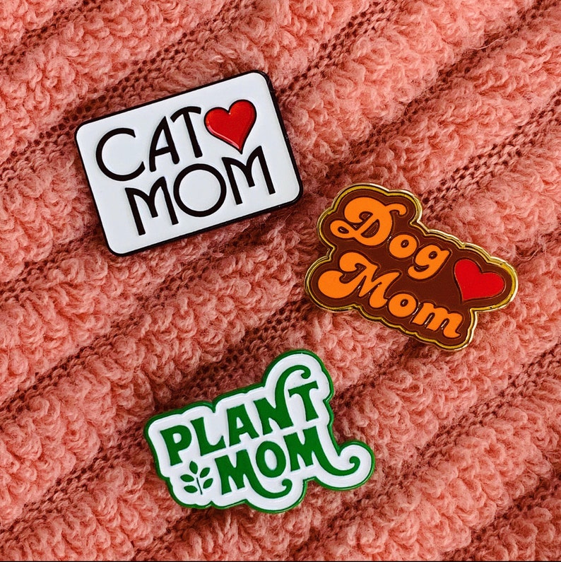 K&ouml;nnte beinhalten: Drei Emaille-Pins im Retro-Design. Die Pins zeigen den Text "Cat Mom", "Dog Mom" und "Plant Mom". Jeder Pin hat ein Herzdesign und ein anderes Farbschema.