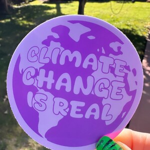May include: A purple round sticker with a white outline. The sticker features a graphic of the Earth with the text "Climate Change is Real" in white bubble letters.