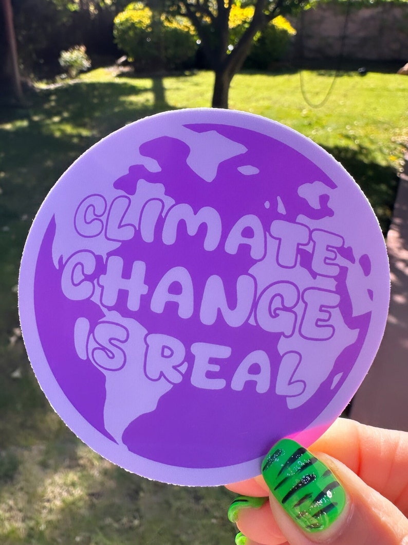 May include: A purple round sticker with a white outline. The sticker features a graphic of the Earth with the text "Climate Change is Real" in white bubble letters.