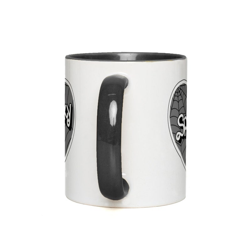 May include: A white ceramic mug with a black rim and handle. The mug has a black and white graphic of a heart with a spiderweb and the word "Spooky" on it.