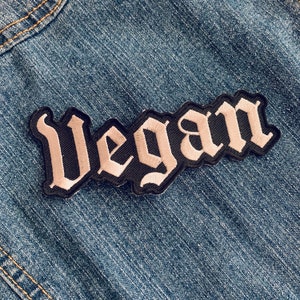 May include: A black and white embroidered patch with the word "Vegan" in a gothic font.