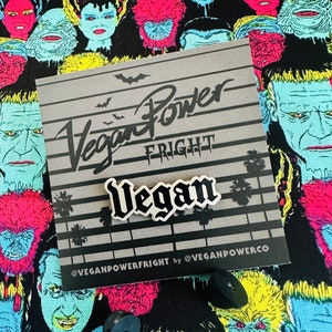 Może przedstawiać: Biała szpilka emaliowana z napisem "Vegan" czarnymi literami. Szpilka znajduje się na szarej kartce z tekstem "Vegan Power Fright" i "@VeganPowerCo".
