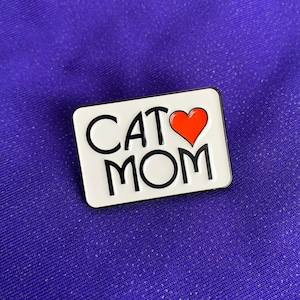 K&ouml;nnte beinhalten: Wei&szlig;er Emaille-Pin mit schwarzem Umriss, mit dem Text "CAT MOM" und einem roten Herzen zwischen den W&ouml;rtern.