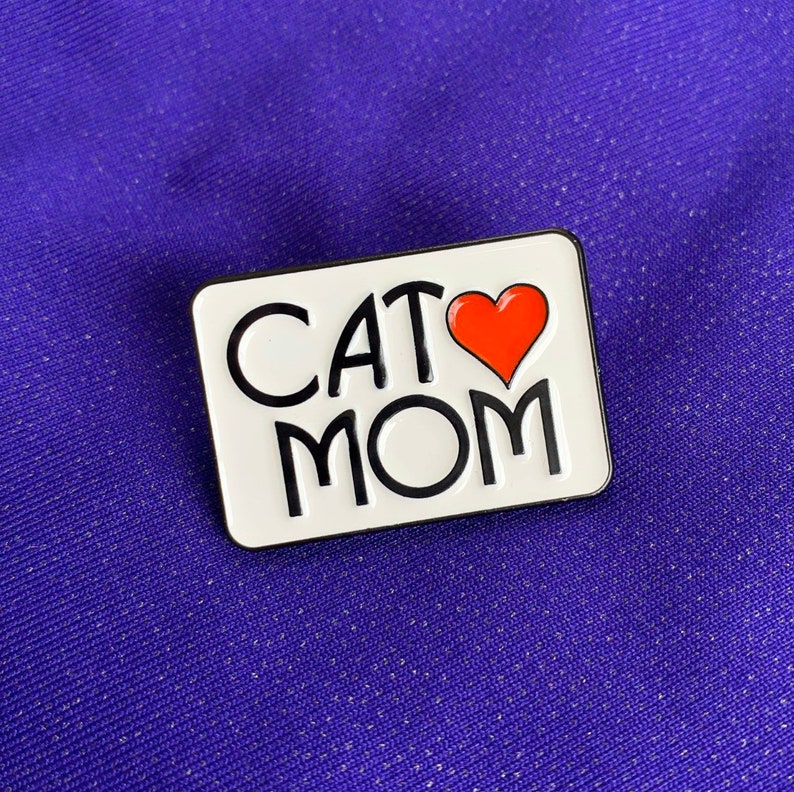 K&ouml;nnte beinhalten: Wei&szlig;er Emaille-Pin mit schwarzem Umriss, mit dem Text "CAT MOM" und einem roten Herzen zwischen den W&ouml;rtern.