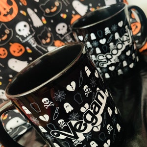 May include: Two black ceramic mugs with white Halloween-themed graphics, including skulls, ghosts, and spiderwebs. One mug says "Vegan" and the other says "Spooky!". A Halloween-themed fabric with pumpkins and ghosts is in the background.