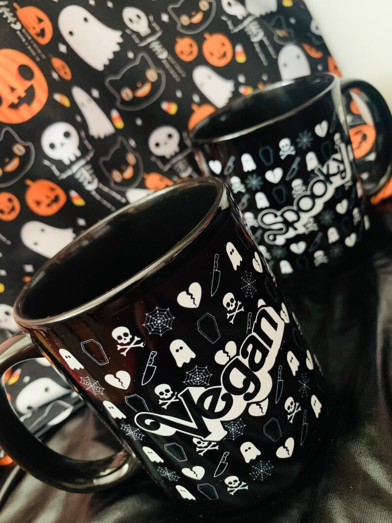 May include: Two black ceramic mugs with white Halloween-themed graphics, including skulls, ghosts, and spiderwebs. One mug says "Vegan" and the other says "Spooky!". A Halloween-themed fabric with pumpkins and ghosts is in the background.