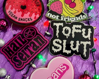 Puede incluir: Una colección de adornos navideños novedosos con varios diseños y texto. Un adorno es una rosquilla rosa con las palabras "HORROR MOVIES VEGAN SNACKS". Otro dice "eat fruit not friends" con ilustraciones de frutas. Otros adornos dicen "Hail Satan", "Tofu Slut" y "Beans".