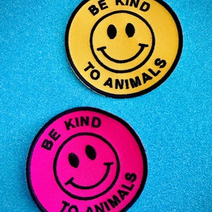 Può includere: Due toppe rotonde da stirare con una faccina sorridente e il testo "Be Kind To Animals". Una toppa è gialla con testo nero e l'altra è rosa con testo nero.