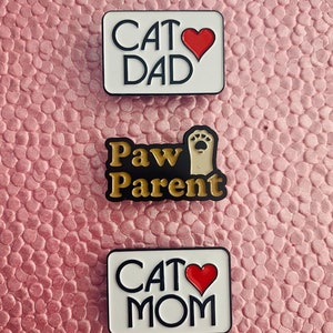 K&ouml;nnte beinhalten: Drei Emaille-Pins mit Katzenmotiv. Der obere Pin ist wei&szlig; mit schwarzem Text, der "Cat Dad" und ein rotes Herz sagt. Der mittlere Pin ist schwarz mit goldenem Text, der "Paw Parent" und eine Pfote sagt. Der untere Pin ist wei&szlig; mit schwarzem Text, der "Cat Mom" und ein rotes Herz sagt.