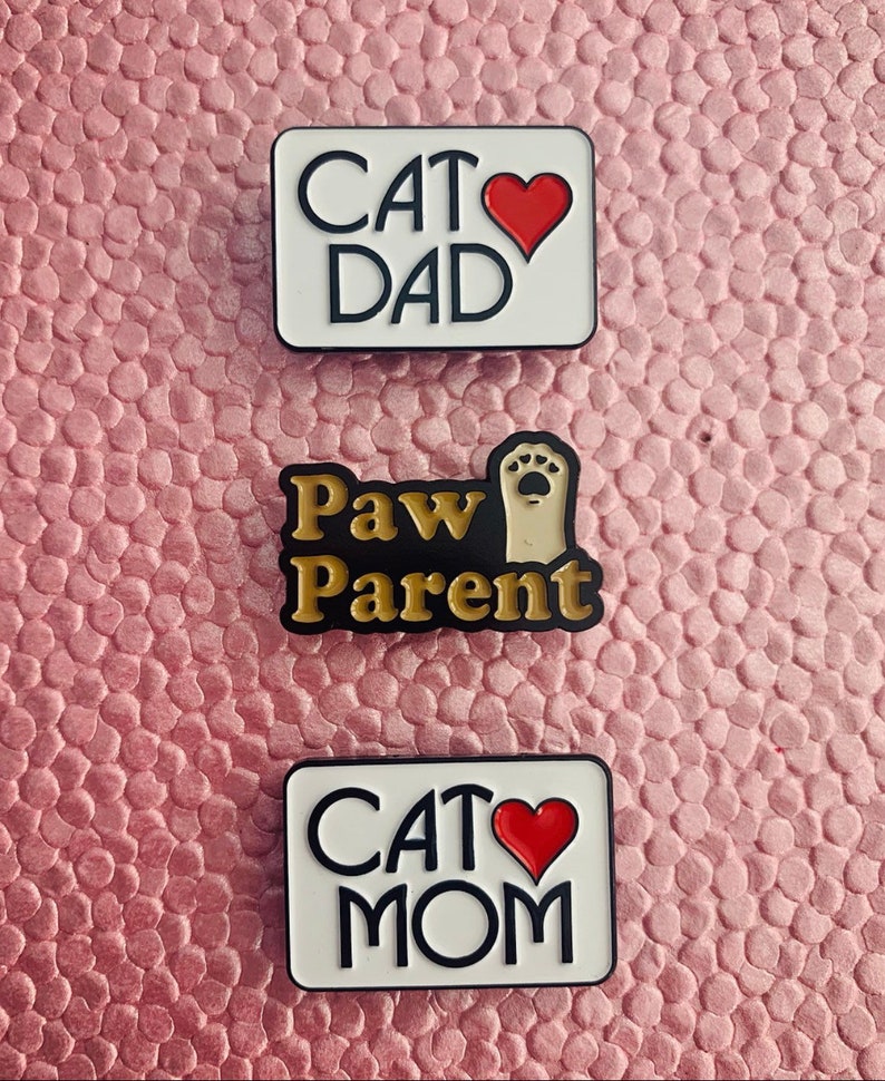 K&ouml;nnte beinhalten: Drei Emaille-Pins mit Katzenmotiv. Der obere Pin ist wei&szlig; mit schwarzem Text, der "Cat Dad" und ein rotes Herz sagt. Der mittlere Pin ist schwarz mit goldenem Text, der "Paw Parent" und eine Pfote sagt. Der untere Pin ist wei&szlig; mit schwarzem Text, der "Cat Mom" und ein rotes Herz sagt.