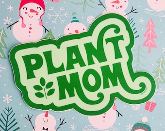 May include: A green sticker with the words "PLANT MOM" in a bold, playful font. The sticker has a light green border and a small leaf design. The background is a light blue pattern with snowmen, snowflakes, and Christmas trees.