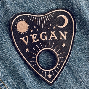 Puede incluir: Parche bordado en blanco y negro con forma de tablero de ouija con la palabra "VEGAN" en el centro. El parche tiene una luna creciente, un sol y estrellas alrededor de los bordes.
