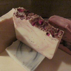 K&ouml;nnte beinhalten: Vier handgemachte Seifen, zwei sind wei&szlig; mit rosa Wirbeln und getrockneten Bl&uuml;tenbl&auml;ttern, eine ist einfarbig hellrosa und eine ist hellgrau mit dunkelgrauen Wirbeln.