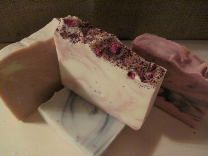 K&ouml;nnte beinhalten: Vier handgemachte Seifen, zwei sind wei&szlig; mit rosa Wirbeln und getrockneten Bl&uuml;tenbl&auml;ttern, eine ist einfarbig hellrosa und eine ist hellgrau mit dunkelgrauen Wirbeln.