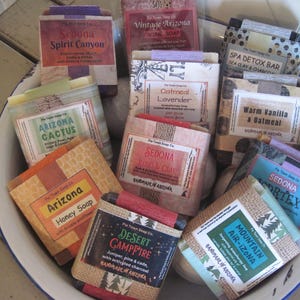 May include: A collection of wrapped soap bars in a white and blue enamel bowl. The soaps have various labels with names like "Arizona Cactus," "Sedona Spirit Canyon," and "Desert Campfire." The labels feature text and decorative elements.