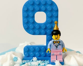 May include: A blue plastic number nine cake topper with a textured brick-like surface pattern. A small plastic figure wearing a gold party hat, a light blue suit jacket with pink accents, a gold bowtie, and pink trousers stands attached to the base.