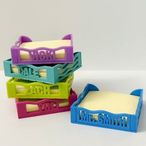 May include: A stack of five colorful plastic note holder dispensers with names "Vicki", "Gale", "Zach", "Yoli", and "Mr. Smith" printed on them.